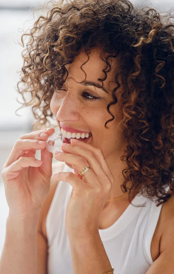 Woman with curly hair smiling while flossing teeth in bright, clean bathroom setting.