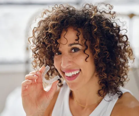 Woman with curly hair brushing teeth, smiling in bright bathroom setting.