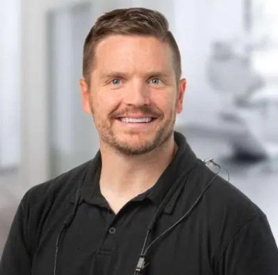 Professional headshot of smiling man in black polo shirt against blurred dental office background.