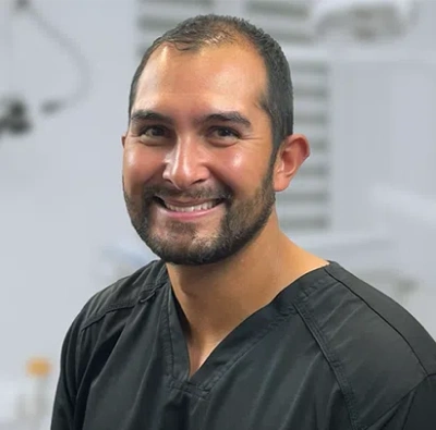 Male dental professional in dark scrubs smiling at camera in clinical setting.