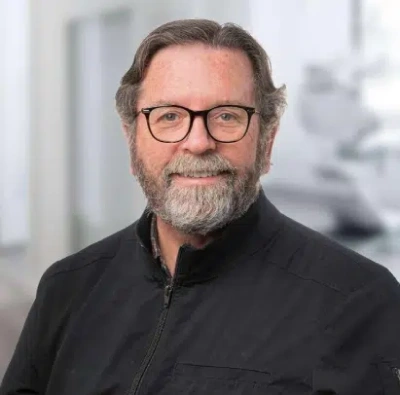 Professional headshot of middle-aged man with glasses and beard in dental office setting.