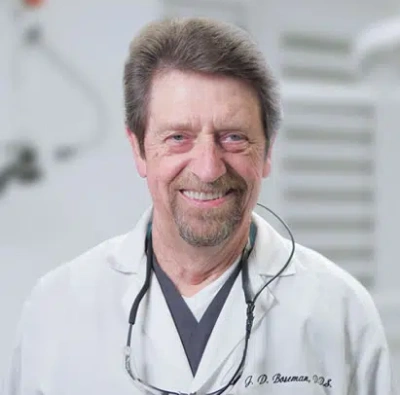 Male dentist smiling at camera wearing white coat and stethoscope in clinical setting.