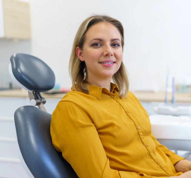Woman in yellow shirt smiling in modern dental office with chair visible behind her.
